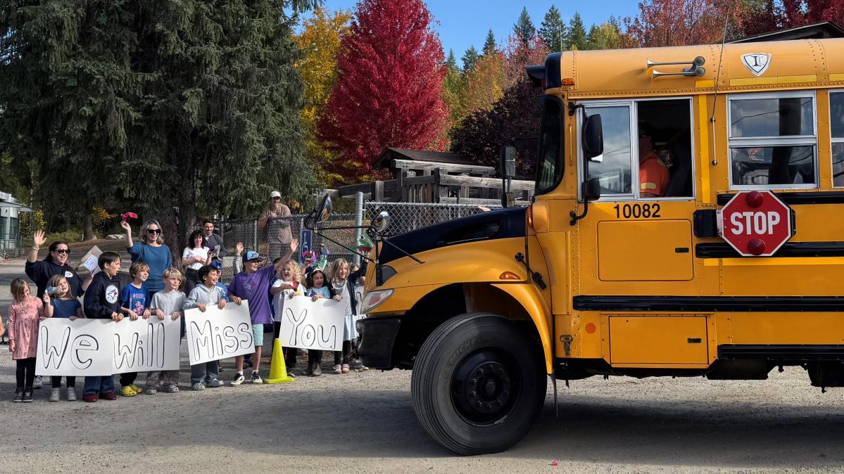 Children hold a sign that says, "We will miss you" while a school bus approaches.