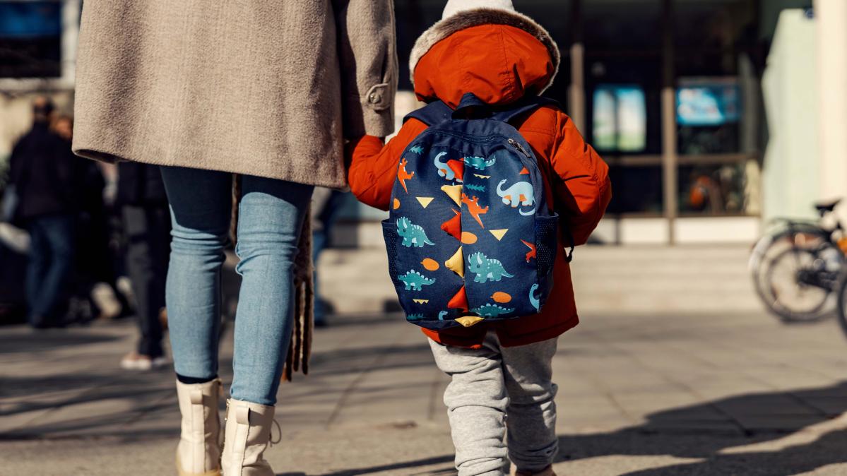 An adult in a beige fall coat walks a child wearing an animal backpack toward a school.