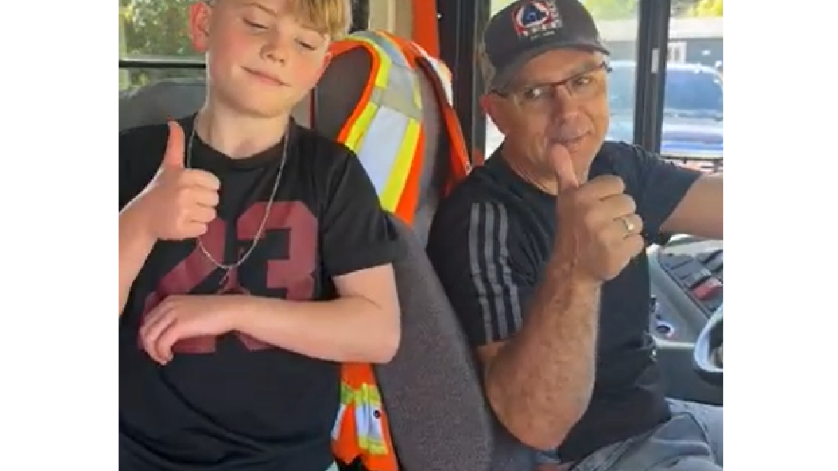 A blonde boy and a bus driver with glasses give a thumbs up inside a school bus