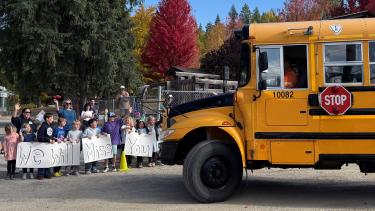Children hold a sign that says, "We will miss you" while a school bus approaches.